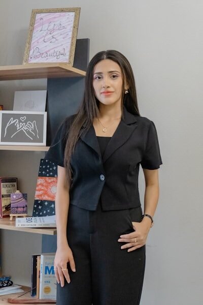 A woman in a black suit stands by a bookshelf.
